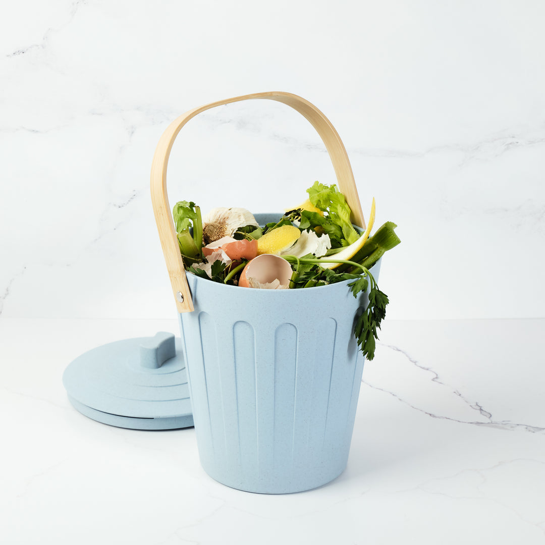 Light blue compost bin with a wooden handle filled with food waste on a white background