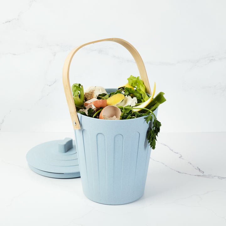 Light blue compost bin with a wooden handle filled with food waste on a white background