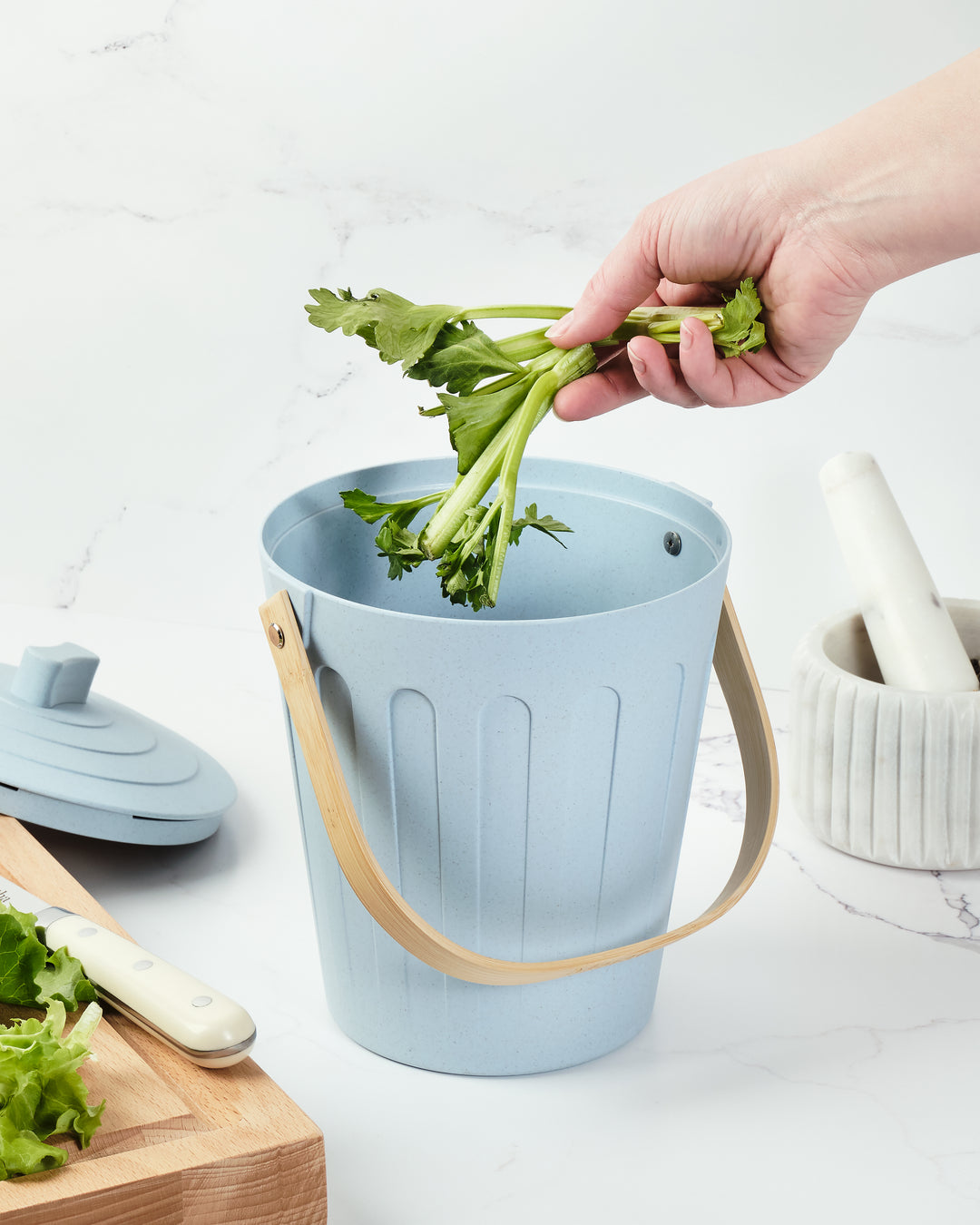 Light blue compost bucket with celery being added, on a white surface with kitchen items.