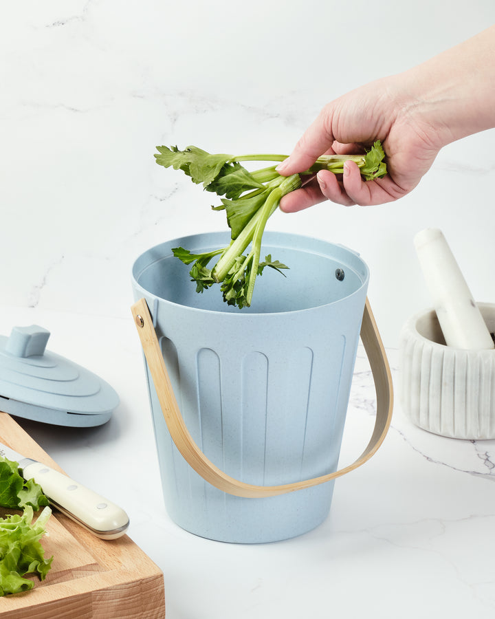Light blue compost bucket with celery being added, on a white surface with kitchen items.